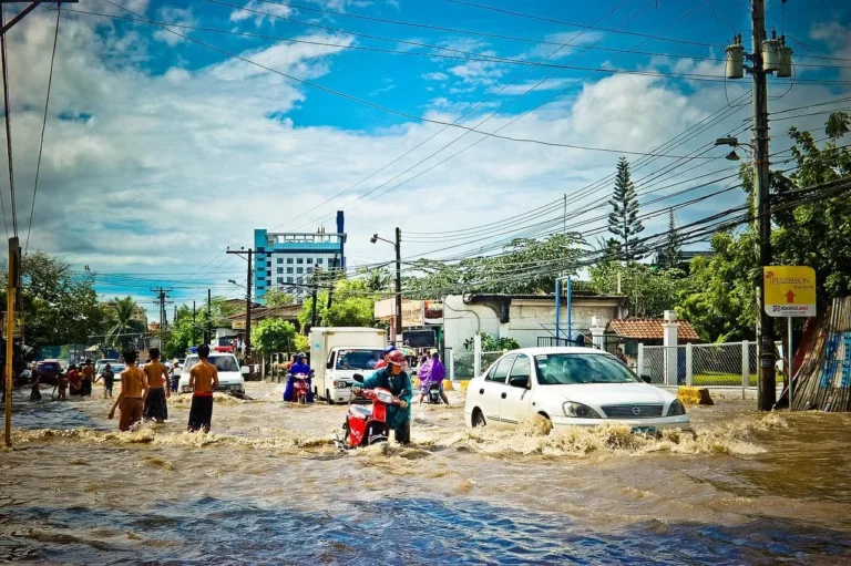 People wade through a street full of muddy running water. Cars are stalled in the street. The water almost reaches the headlights.