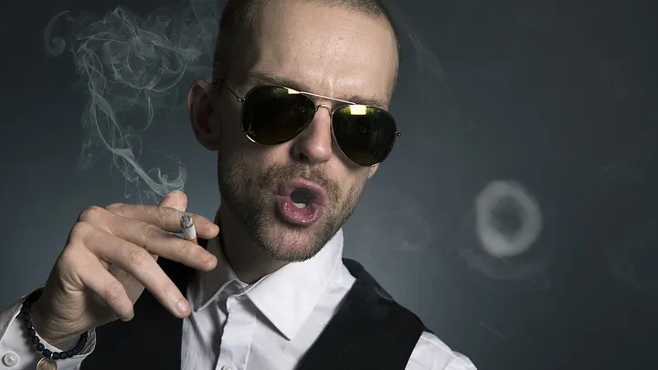 Man in a collared shirt, fancy vest, and sunglasses, holding a cigarette, blowing smoke rings.