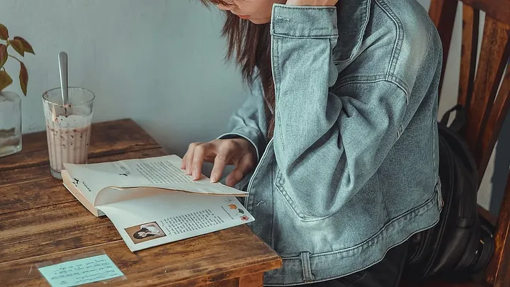 Long-haired person in a denim jacket reading a book at a table with a milkshake in a glass