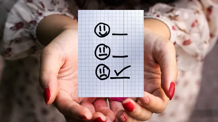 Person with red-painted nails holds a piece of graph paper with crudely hand-drawn smiley, flat-faced, and frowney faces. The line next to the frowney face is checked off.