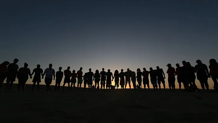 Silhouettes of lots of people standing in a field against the glowing horizon.