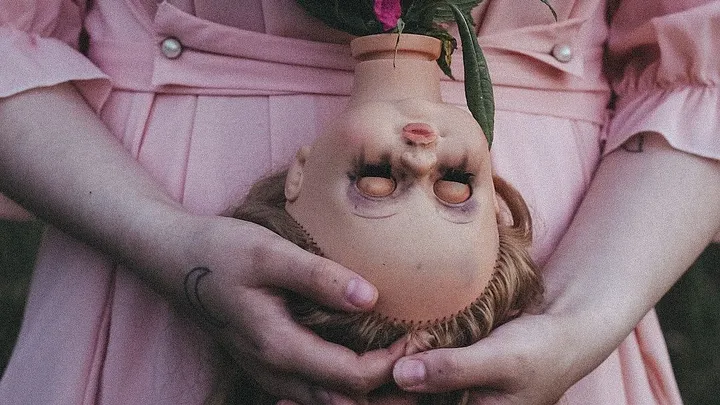 person wearing pink dress holds a decapitated doll head upside down in her palms