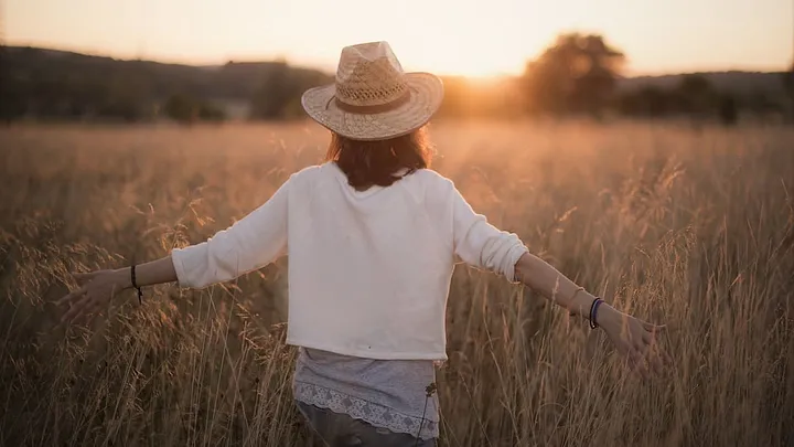 person with long hair and sunset, wearing lace under a white shirt, raising hands in a field of tall grass glowing with sunset