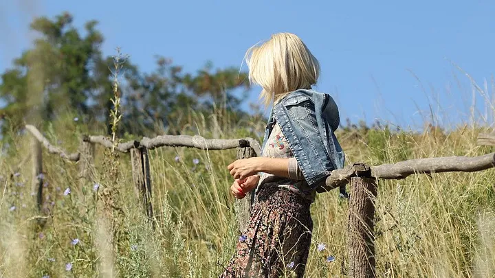 A person with shoulder-length blonde hair, wearing a denim jacket and a skirt, standing alone in a field of long grass.