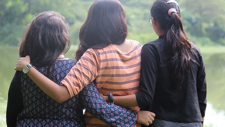 three friends with long dark hair, seen from behind, strolling with their arms around each other