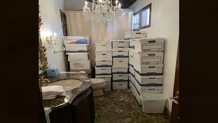 Cardboard boxes in a bathroom. The sink is visible. The boxes block access to the shower, which has a white curtain. A glass chandelier hangs from the ceiling.