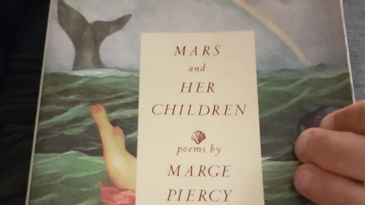 holding a paperback of Mars and Her Children by Marge Piercy