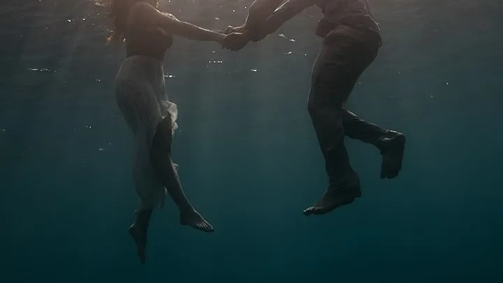 Two indistinct bodies, one more feminine and one more masculine, floating underwater, holding hands