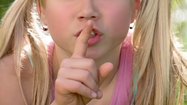 small child in blond pigtails and earrings puts her finger to her lips as if to shush someone