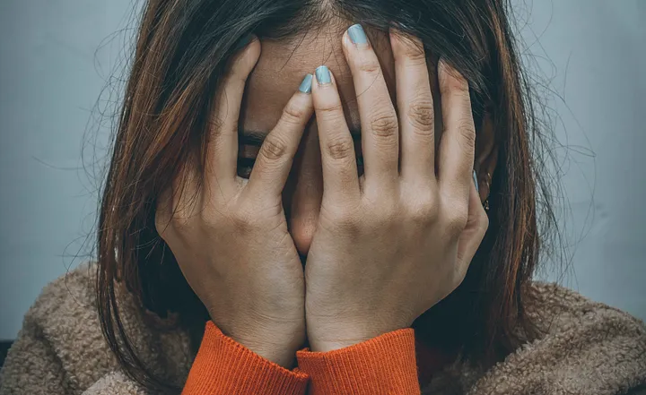 A person with long hair and painted blue nails hides her face in her hands as if in frustration or shame.