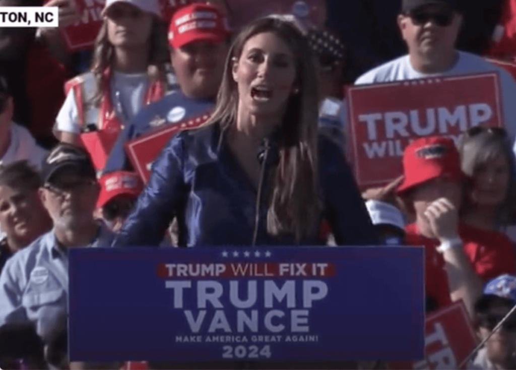 Alina Habba yelling into a microphone at a rally behind a Trump/Vance platform