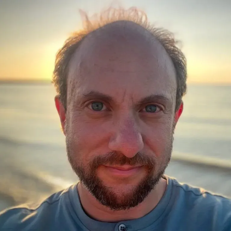 headshot of Tucker Lieberman gazing into camera, hazy ocean in the background. He's a 40-something white man, balding and with a beard, wearing a blue T-shirt.