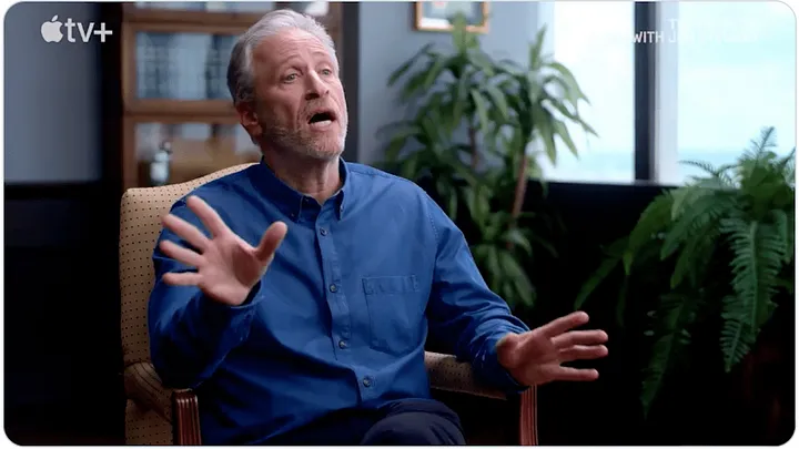 Jon Stewart, seated in a living room, moving his hands animatedly, talking