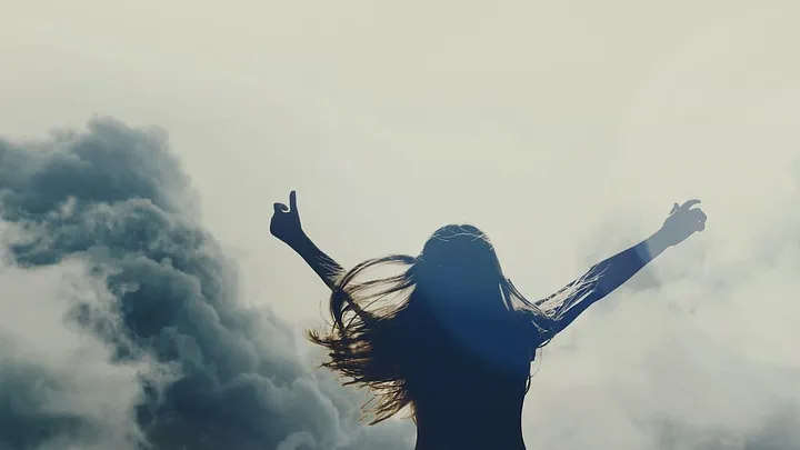 silhouette of long-haired person lifts arms to the sky. there's a dramatic cloud.