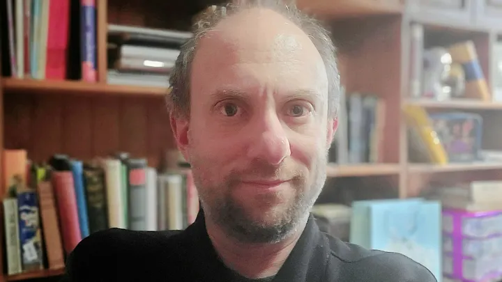 photo of Tucker Lieberman in front of a colorful bookshelf. He's a balding white Jewish man with whiskers, wearing a black jacket, smiling slightly