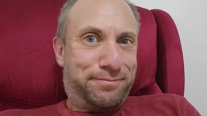 Tucker Lieberman seated in a wine-colored armchair, wearing a similarly colored T-shirt. He's in his 40s, white, blue-eyed, balding and bearded. He has a quizzical expression.