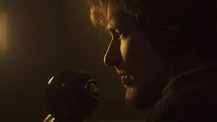pensive-looking young bearded person in a dark room with mouth close to a large microphone