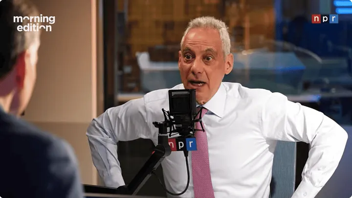 video still of NPR Morning Edition interview. Rahm Emanuel has gray hair and is wearing a collared white shirt and pink tie. He's speaking into a microphone, his eyes wide, his hands on his hips.