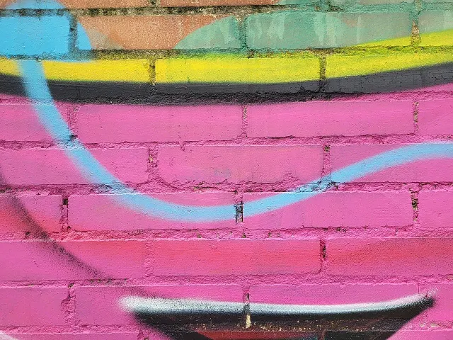 brick wall painted with abstract graffiti in bright '90s colors, mostly bright pink with a little blue and yellow. there appears to be a triangle at the bottom.
