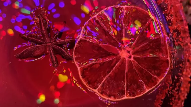 closeup of a cocktail. a thin lime slice and anise star floating in a red drink with colored lights reflecting on the surface
